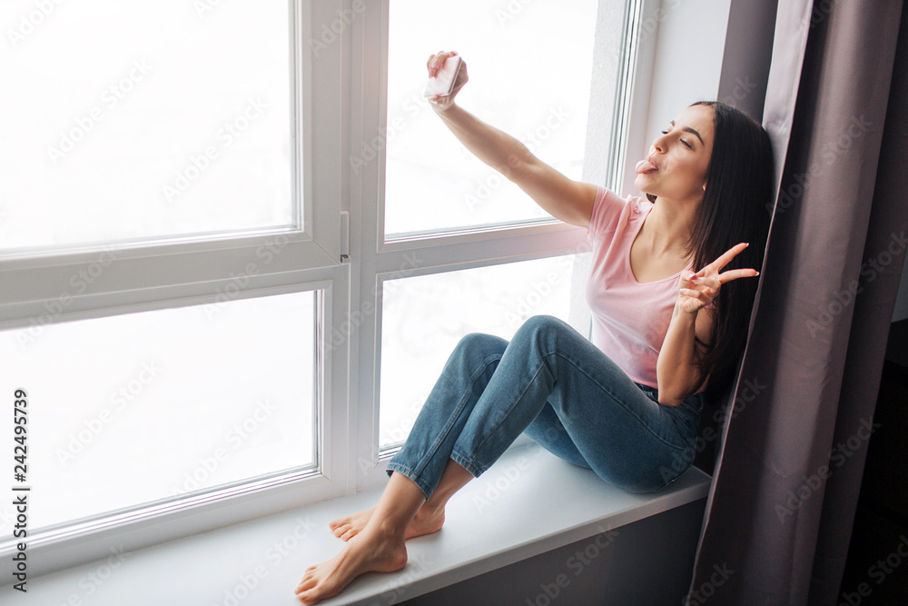 Cheerful young woman take selfie at window. She smile and pose. Model show piece sign with fingers. Cheerful young woman take selfie at window. She smile and pose. Model show piece sign with fingers.