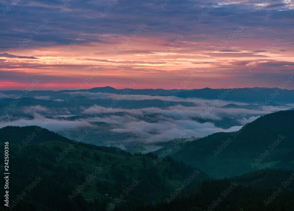 Panoramic photo of Mountain range through the morning colorful fog in the summer. Beautiful sunrise in the Carpathian Mountains with visible silhouettes and pink clouds in the sun rays.