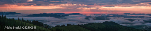 Panoramic photo of Mountain range through the morning colorful fog in the summer. Beautiful sunrise in the Carpathian Mountains with visible silhouettes and pink clouds in the sun rays.