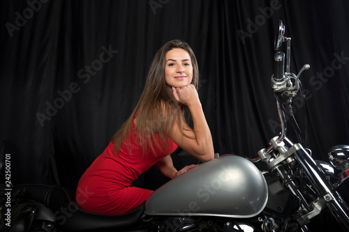 Beautiful young brunette in bright red dress sitting on a chrome motorcycle on a black background