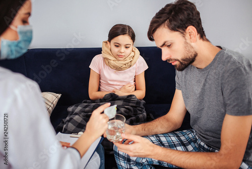 Small girl sit on sofa. her legs and throat are covered. She look how doctor pour in powder medicine into glass of water. Young man help. They look concentrated.