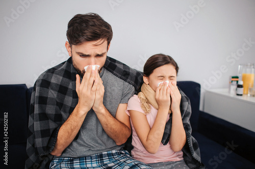 Young man sit with small girl on bed and sneezing into white tissues. They caught cold. Family covered with blanket.