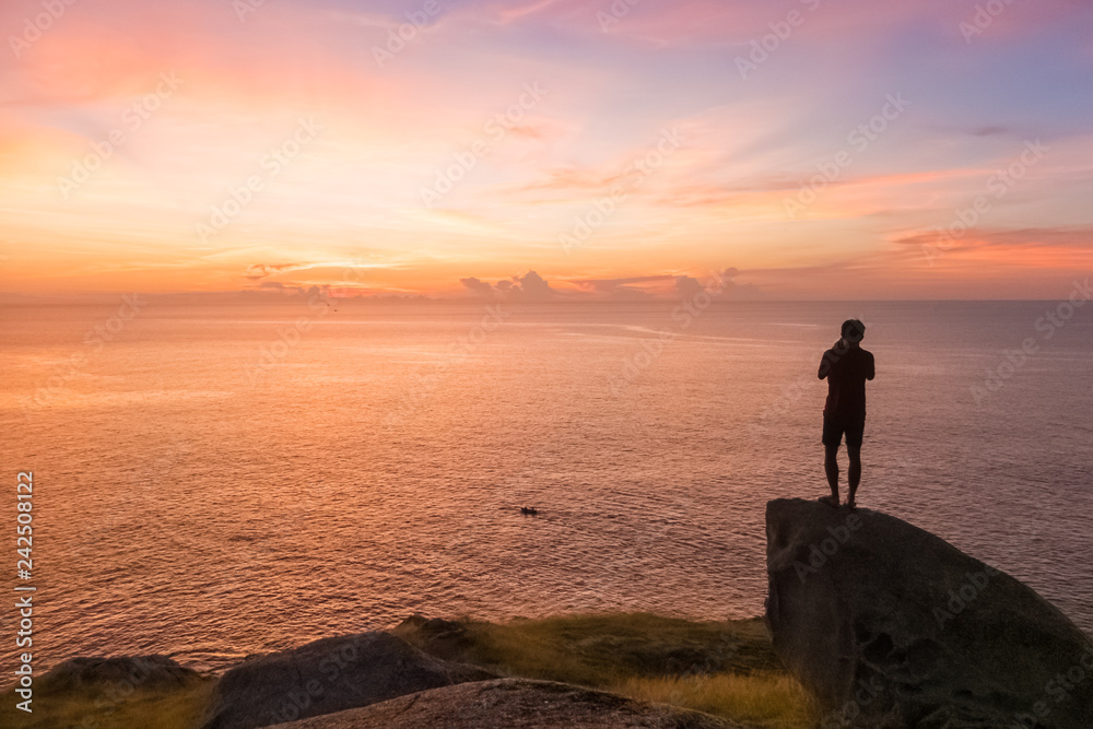 silhouette of young man stands on the stone and takes a photo with small boat and beautiful amazing sea sunset of orange red colors silhouette of young man stands on the stone and takes a photo with small boat and beautiful amazing sea sunset of orange red colors