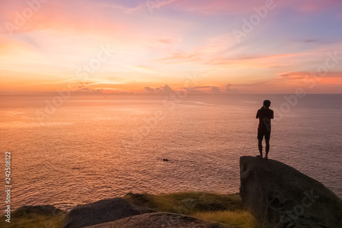 silhouette of young man stands on the stone and takes a photo with small boat and beautiful amazing sea sunset of orange red colors