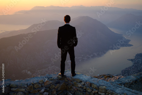  back of a young man in costume standing on a mountain at sunset. The groom or businessman looks from the top. © Boyarkina Marina