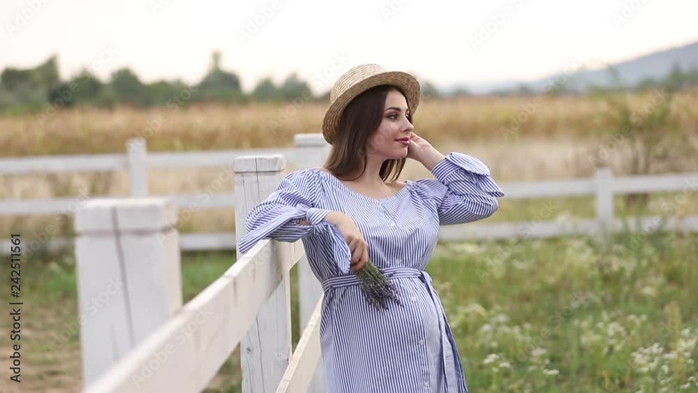 Pregnant woman in nature try on knitted hat. Background of field and white fance near the farm