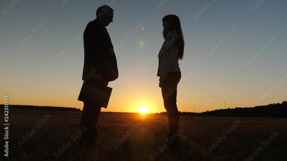 silhouette businessman and young business woman made deal, pass black briefcase to each other and shake hands against backdrop of sunset against blue sky