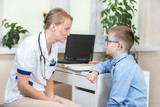 Lekarka w białym kitlu i stetoskopem na szyi siedzi w gabinecie lekarskim. Rozmawia z chłopcem w niebieskiej koszuli i okularach. 