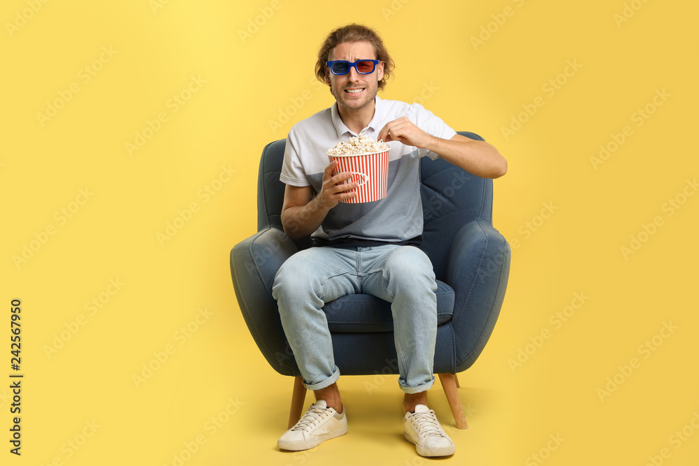 Emotional man with 3D glasses and popcorn sitting in armchair during cinema show on color background Emotional man with 3D glasses and popcorn sitting in armchair during cinema show on color background