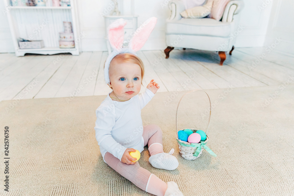 Little child girl wearing bunny ears on Easter day. Girl holding basket with painted eggs sitting on floor at home, having fun on Easter egg hunt. Happy Easter holiday celebration spring concept Little child girl wearing bunny ears on Easter day. Girl holding basket with painted eggs sitting on floor at home, having fun on Easter egg hunt. Happy Easter holiday celebration spring concept