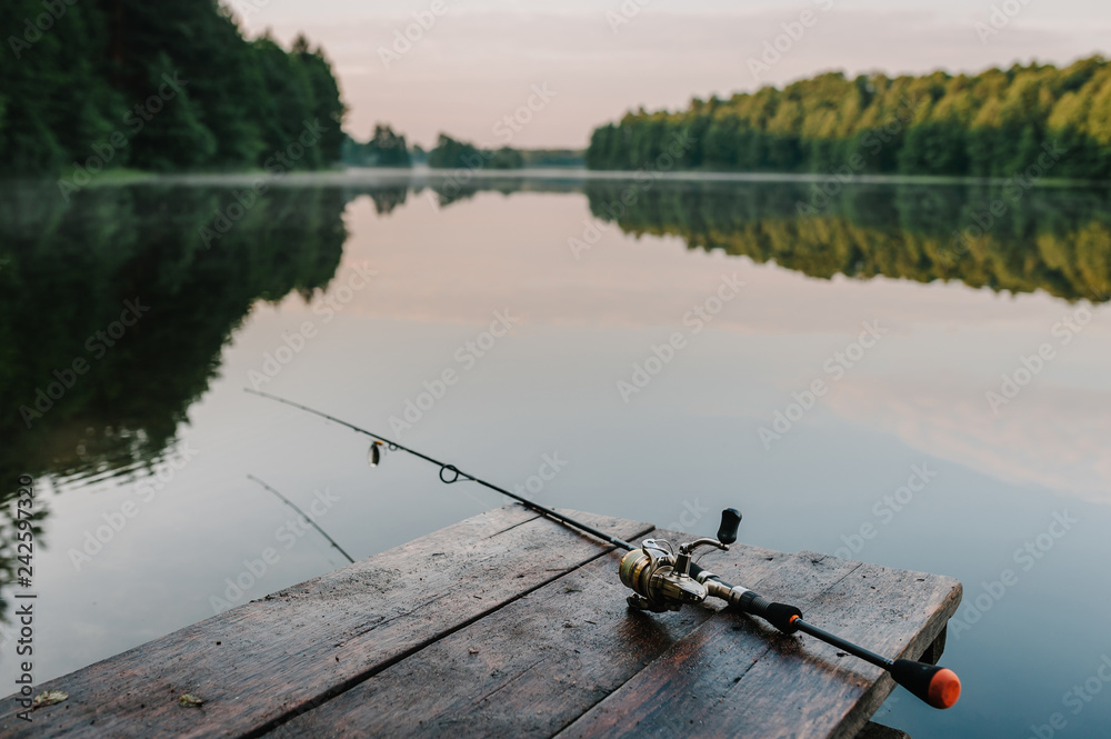 Fishing rod, spinning reel on the background pier river bank. Sunrise. Fog  against the backdrop of lake. Misty morning. wild nature. The concept of  rural getaway. Article about fishing day. Stock Photo