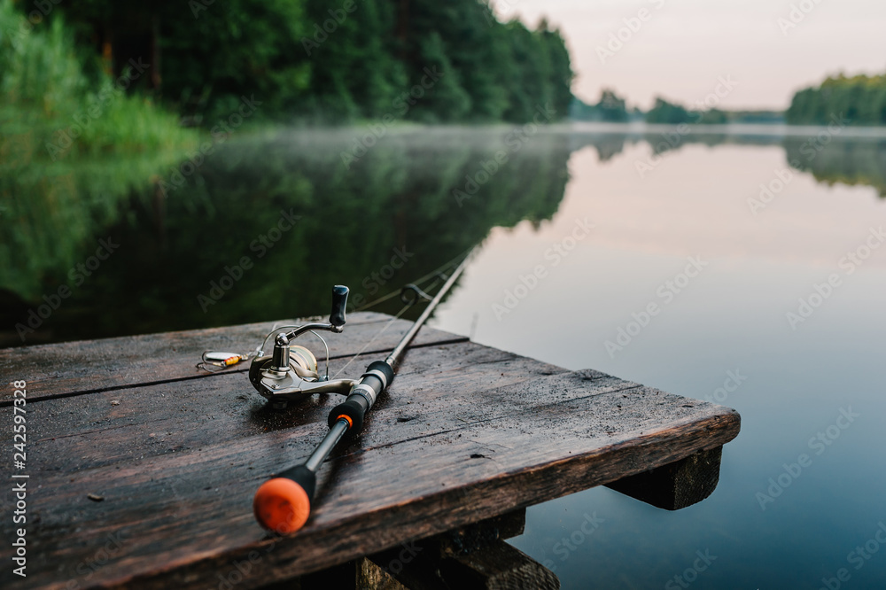 Fishing rod, spinning reel on the background pier river bank. Sunrise. Fog  against the backdrop of lake. Misty morning. wild nature. The concept of  rural getaway. Article about fishing day. Photos