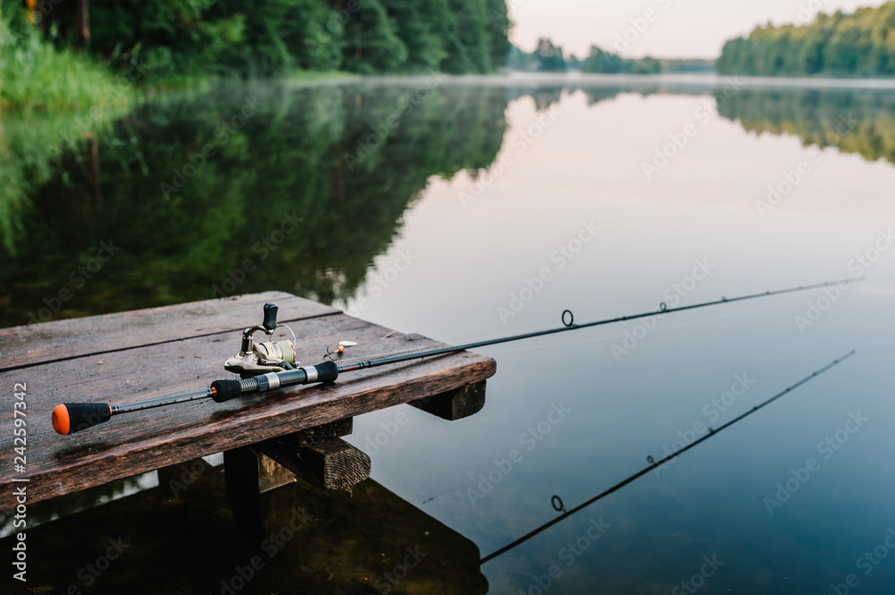 Fishing rod, spinning reel on the background pier river bank. Sunrise. Fog  against the backdrop of lake. Misty morning. wild nature. The concept of  rural getaway. Article about fishing day. Stock Photo