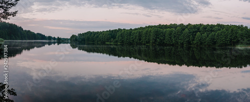 Lake with spring trees panorama photo. Tranquil landscape at a lake, with the vibrant blue sky, white clouds and the trees reflected symmetrically in the clean blue water. © Serhii