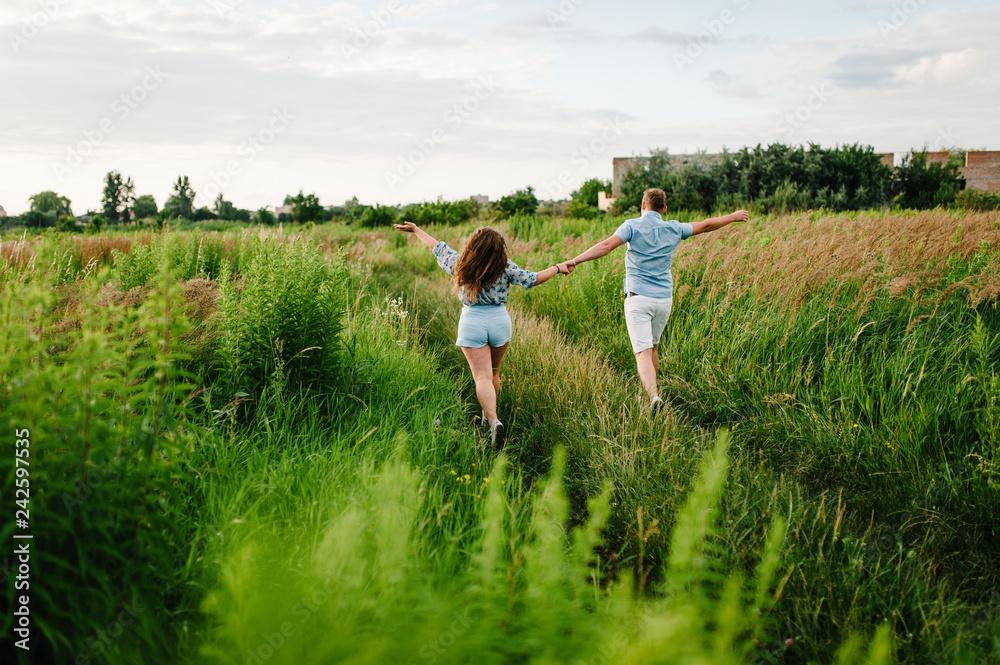 Rear view of a romantic man and woman walking on field grass, nature enjoying stunning sunset. Concept of lovely family holding hands. Young couple running and looking away. Rear view of a romantic man and woman walking on field grass, nature enjoying stunning sunset. Concept of lovely family holding hands. Young couple running and looking away.