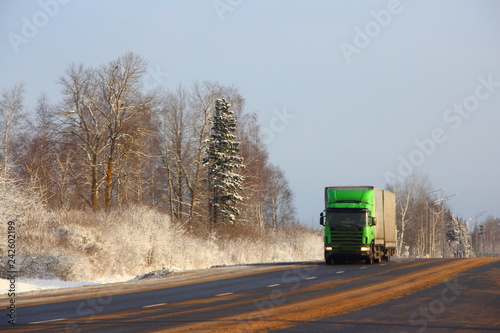 European winter road landscape - two lane asphalt road with green truck front view and snowy trees background