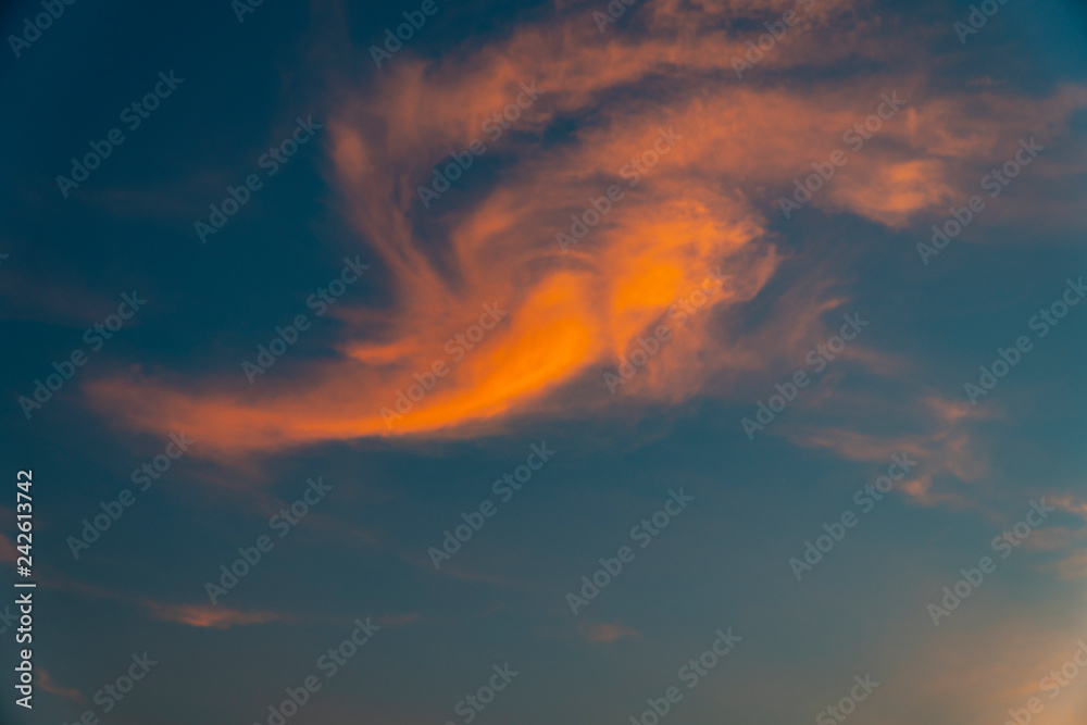 Blue sky with a reflection of the light of sunset at dusk on the cloudy shapes that look like an angel in orange color that is flying in the air.