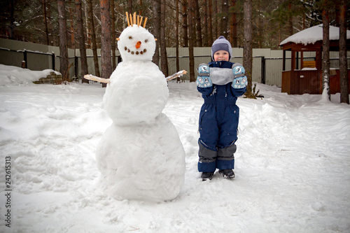 A little boy in the winter in the garden stands next to a snowman and looks at the camera smiling