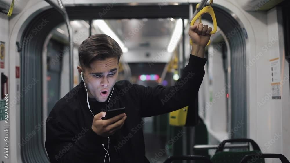 Happy businessman cheering celebrating looking at smartphone. Young urban professional successful business man receiving good news riding in public transport. He holds the handrail
