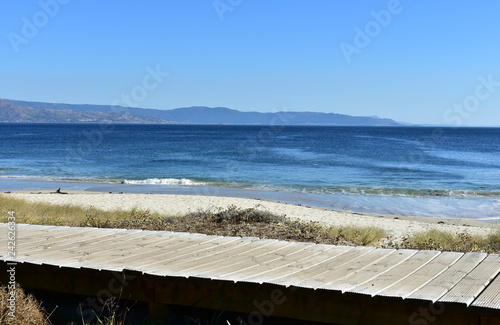 Beach in a bay with wooden boardwalk and vegetation in sand dunes. Blue sea with small waves and clear sky. Sunny day. Galicia  Spain.