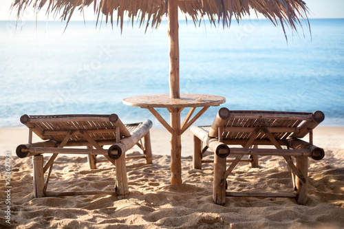 Fototapeta Naklejka Na Ścianę i Meble -  Two empty bamboo loungers and table under straw umbrella on white sand lonely beach, blue sea background in the morning sun light, nobody, design for tourist poster, travel banner, relax vacation card