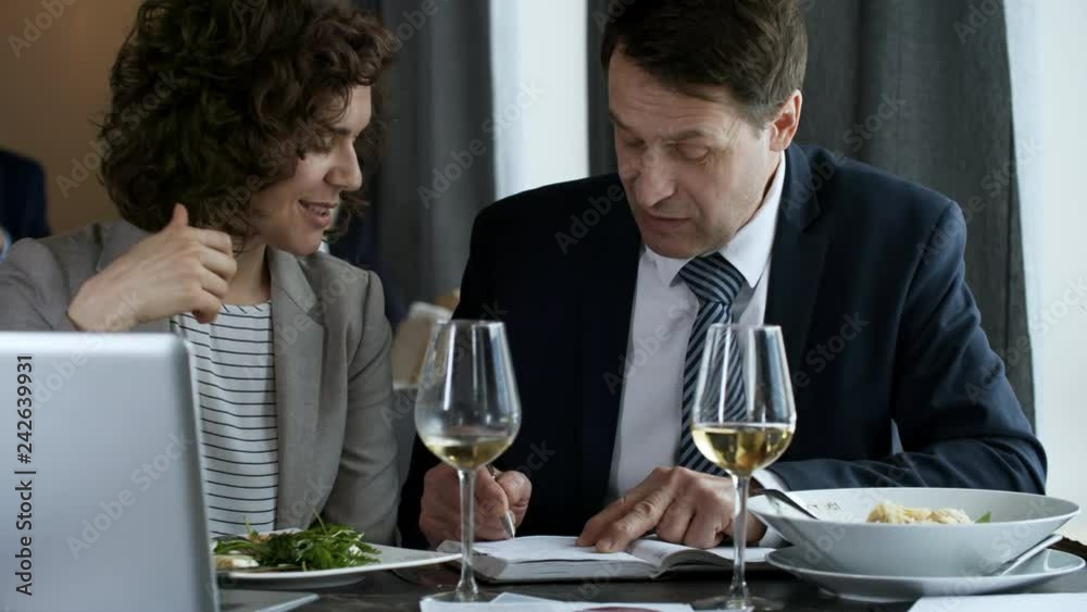 Mid-aged businessman and young businesswoman writing down plans in notepad and discussing them while having lunch in the restaurant. There are dishes, wine glasses and laptop on table Mid-aged businessman and young businesswoman writing down plans in notepad and discussing them while having lunch in the restaurant. There are dishes, wine glasses and laptop on table