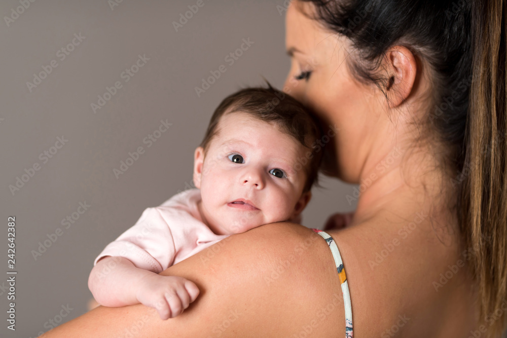Closeup photo of a baby while her mother holding her on her chest while standing in front of a grey background in a studio. Closeup photo of a baby while her mother holding her on her chest while standing in front of a grey background in a studio.