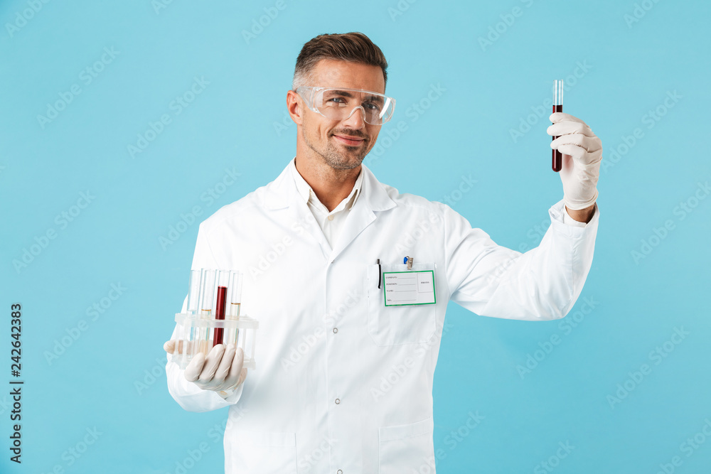 Portrait of happy medical doctor wearing glasses holding test tubes with blood, standing isolated over blue background Portrait of happy medical doctor wearing glasses holding test tubes with blood, standing isolated over blue background