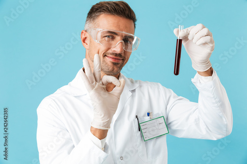 Portrait of medical worker wearing white uniform holding test tubes with blood, standing isolated over blue background