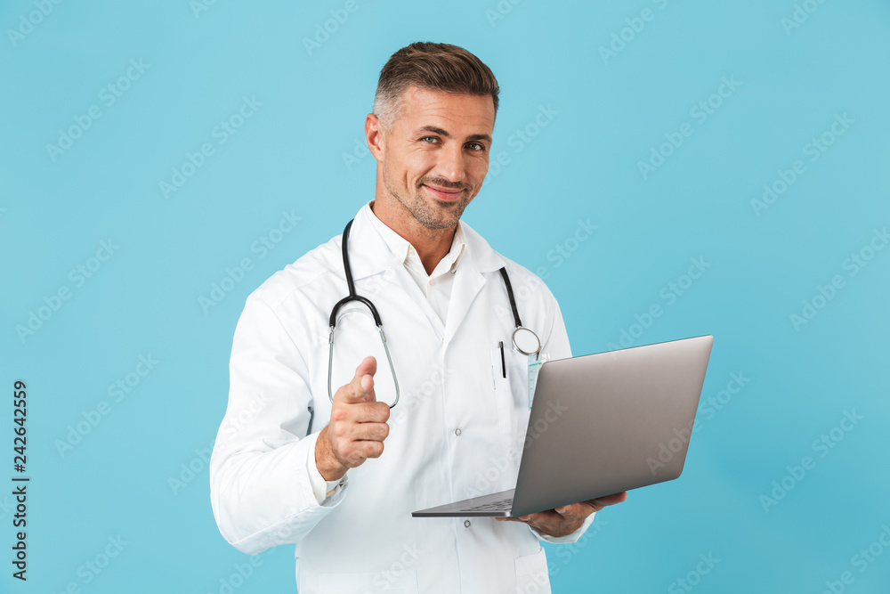Photo of handsome man wearing white medical coat and stethoscope holding laptop, standing isolated over blue background Photo of handsome man wearing white medical coat and stethoscope holding laptop, standing isolated over blue background