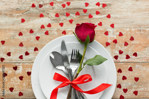 valentines day and romantic dinner concept - close up of red rose flower on set of dishes with cutlery and hearts on wooden table