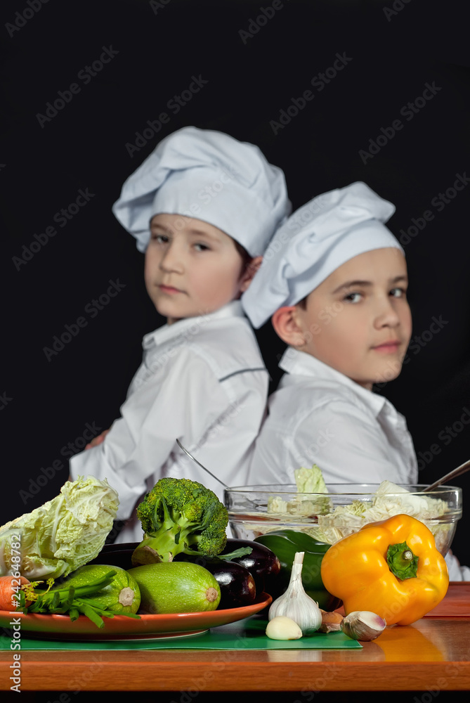 The boys in the cooks costumes cut the salad in the kitchen. Children prepare healthy food. Friendly cooperation of cooks. Isolated on black background. The boys in the cooks costumes cut the salad in the kitchen. Children prepare healthy food. Friendly cooperation of cooks. Isolated on black background.