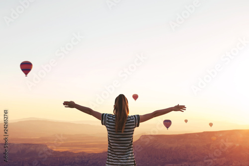 A girl stands on top of a mountain in solitude, admires the beautiful view of the natural landscape and air balloons in Cappadocia in Turkey and raises her hands up showing how she is free and happy