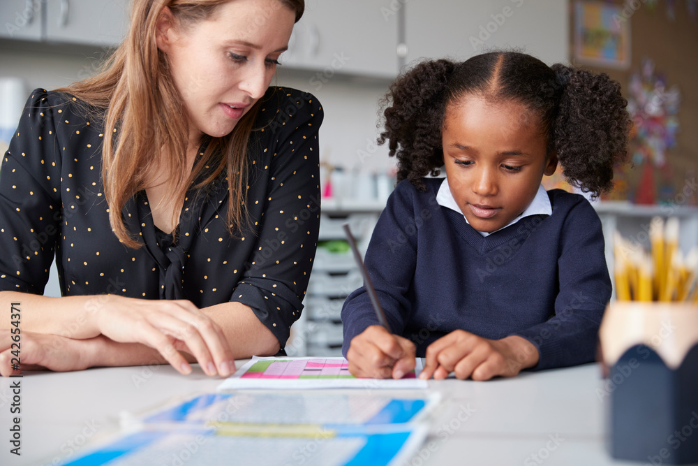 Young female primary school teacher working one on one with a schoolgirl watching her write at a table in a classroom, close up Young female primary school teacher working one on one with a schoolgirl watching her write at a table in a classroom, close up