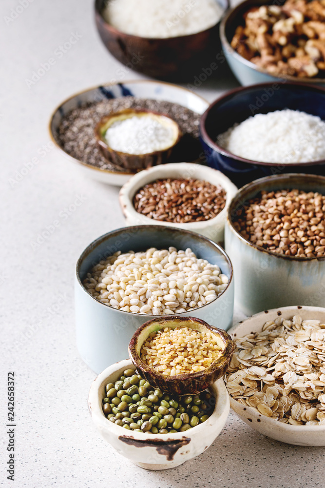 Variety of raw uncooked grains superfood cereal chia seeds, linen, sesame, mung bean, walnuts, tapioca, wheat, buckwheat, oatmeal, coconut, rice in ceramic bowls over grey spotted background. Variety of raw uncooked grains superfood cereal chia seeds, linen, sesame, mung bean, walnuts, tapioca, wheat, buckwheat, oatmeal, coconut, rice in ceramic bowls over grey spotted background.