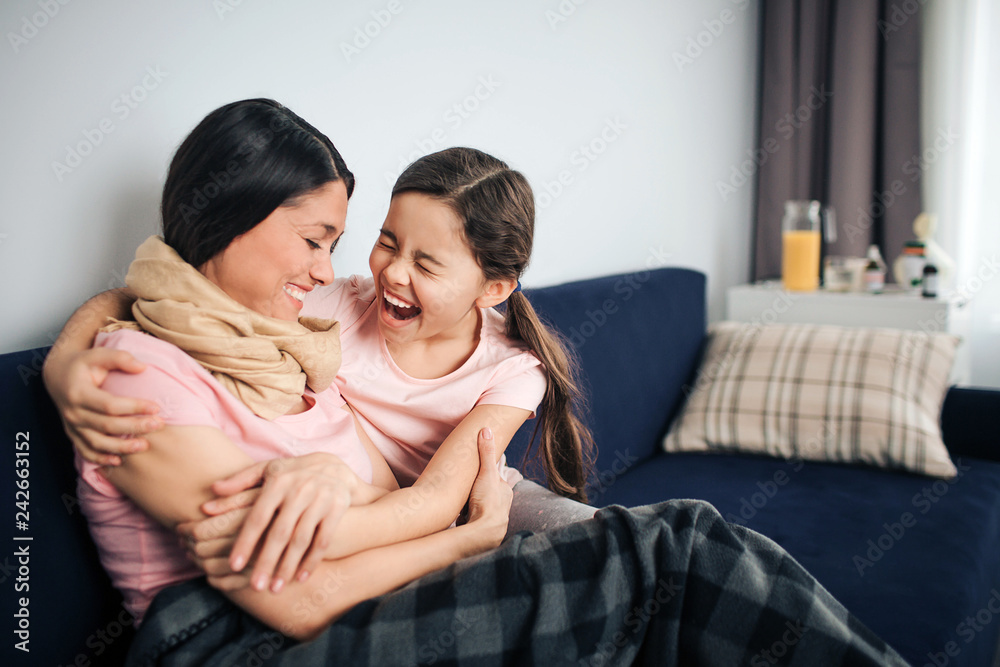 Happy and positive young woman sit together with her daughter on couch in room. They laugh out loud. Young woman is sick. Her throat is wrapped with scarf. Girl embrace mother. Happy and positive young woman sit together with her daughter on couch in room. They laugh out loud. Young woman is sick. Her throat is wrapped with scarf. Girl embrace mother.
