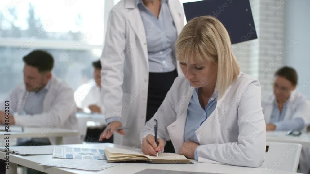 Middle aged blonde woman in lab coat writing down at desk while taking a test on training course for healthcare specialists; female doctor with clipboard walking in seminar room and helping students Middle aged blonde woman in lab coat writing down at desk while taking a test on training course for healthcare specialists; female doctor with clipboard walking in seminar room and helping students