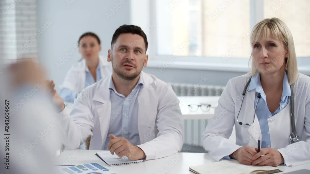 Male and female practitioners in lab coats sitting at desk on health seminar and talking with coach Male and female practitioners in lab coats sitting at desk on health seminar and talking with coach