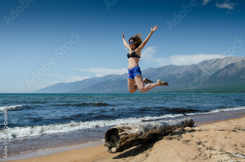 Girl jumping from very old wooden rotten log aground on the beach, near lake Baikal, Russia, on a sunny day in the summer