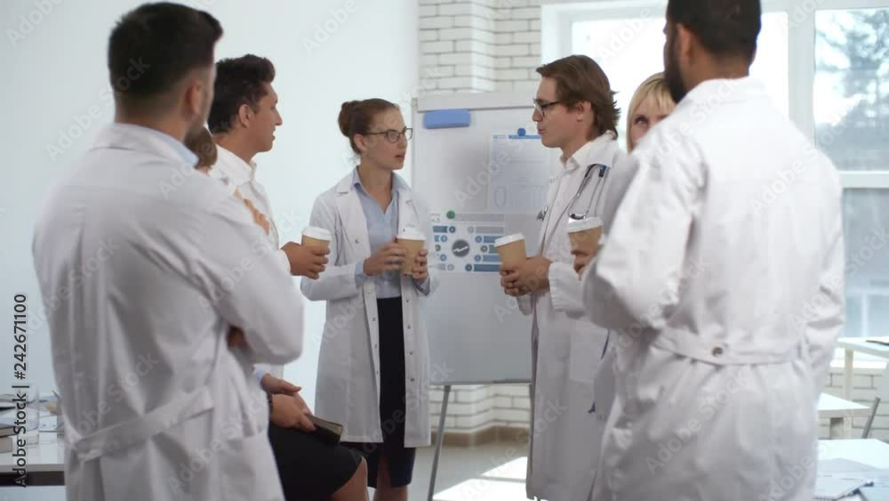 Beautiful female doctor in lab coat standing by flipchart, holding coffee cup and discussing seminar with group of mixed raced colleagues during break Beautiful female doctor in lab coat standing by flipchart, holding coffee cup and discussing seminar with group of mixed raced colleagues during break