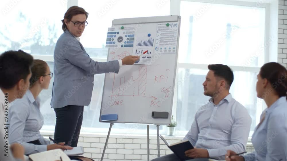 Male CEO using flipchart while giving presentation to colleagues sitting in circle and writing down in notepads during corporate training