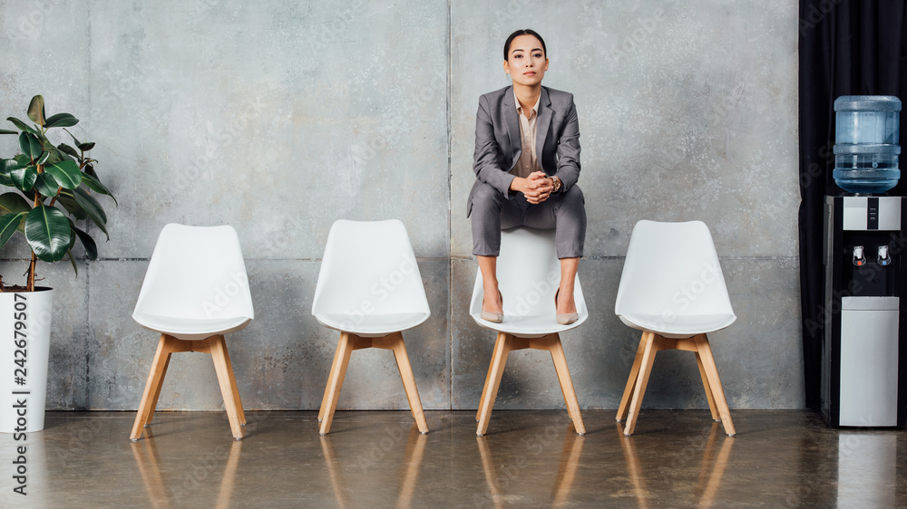Fototapeta premium serious asian businesswoman in formal wear sitting on chair and looking at camera in waiting hall