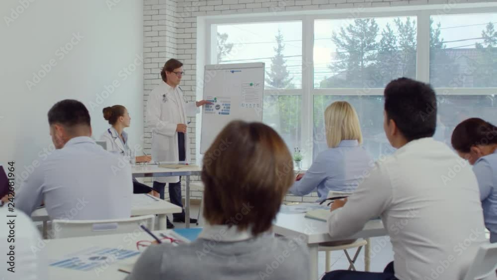 Experienced male doctor in lab coat explaining something on flipchart and giving presentation to healthcare workers in seminar room while his female assistant with stethoscope writing down at desk Experienced male doctor in lab coat explaining something on flipchart and giving presentation to healthcare workers in seminar room while his female assistant with stethoscope writing down at desk