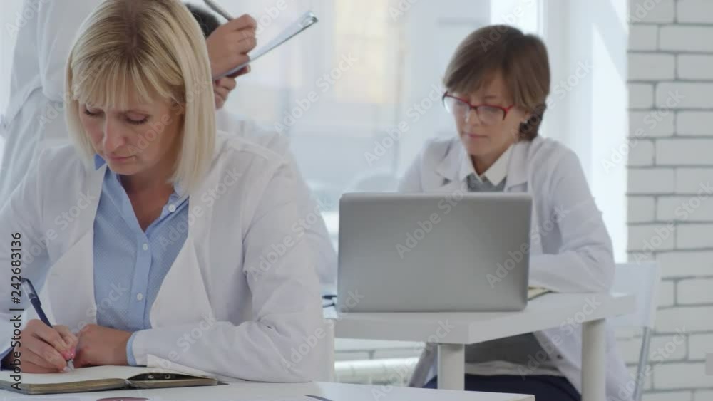 Tracking shot of male and female doctors in lab coats taking test during educational seminar and writing down at desks while female coach walking with clipboard and helping them Tracking shot of male and female doctors in lab coats taking test during educational seminar and writing down at desks while female coach walking with clipboard and helping them