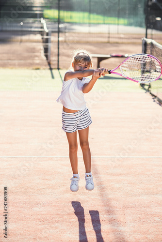 Female little tennis player in white sport uniform practice in hitting with tennis racket at the training on outdoor court on the sunset background. © gorynvd