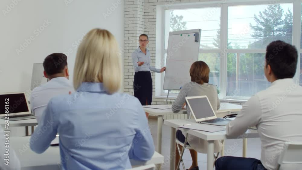 Young businesswoman drawing diagram on flipchart while female office worker raising arms and asking a question during corporate education course Young businesswoman drawing diagram on flipchart while female office worker raising arms and asking a question during corporate education course