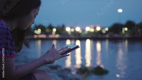 Attractive asian woman using digital tablet during sitting near the river at night with street lights reflected on water.