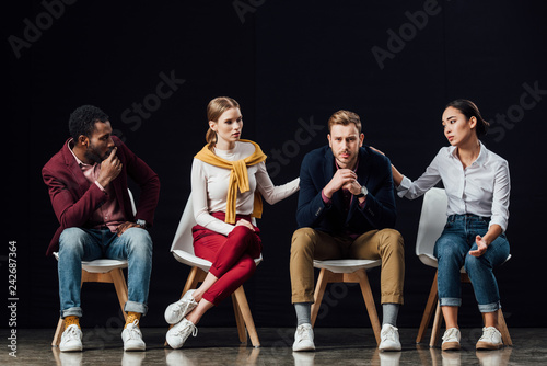multiethnic group of people sitting on chairs and consoling upset man isolated on black