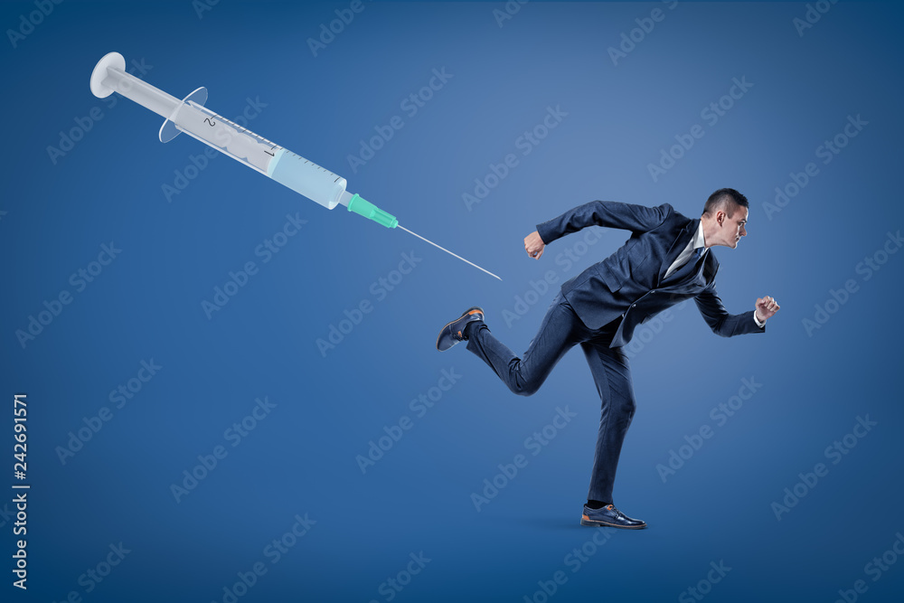 A side view of a businessman running from a huge syringe floating in the air on a blue background. A side view of a businessman running from a huge syringe floating in the air on a blue background.