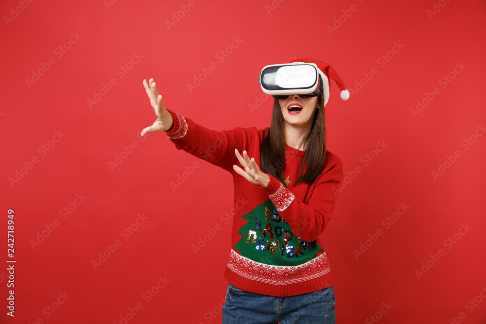 Young Santa girl looking in headset touch something like push click on button, pointing at floating virtual screen isolated on red background. Happy New Year 2019 celebration holiday party concept.